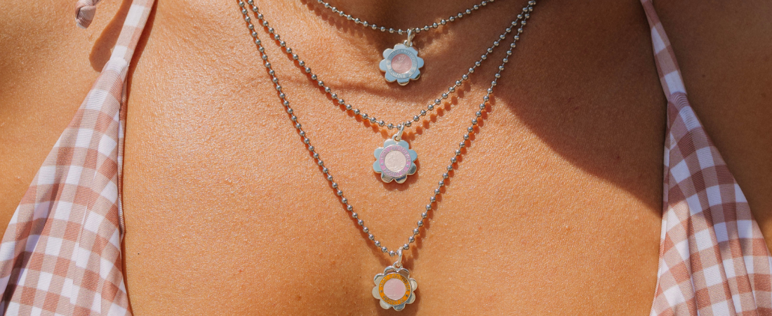 Silver flower charm necklace with pastel enamel Saint Christopher medallions worn on a sunlit collarbone
